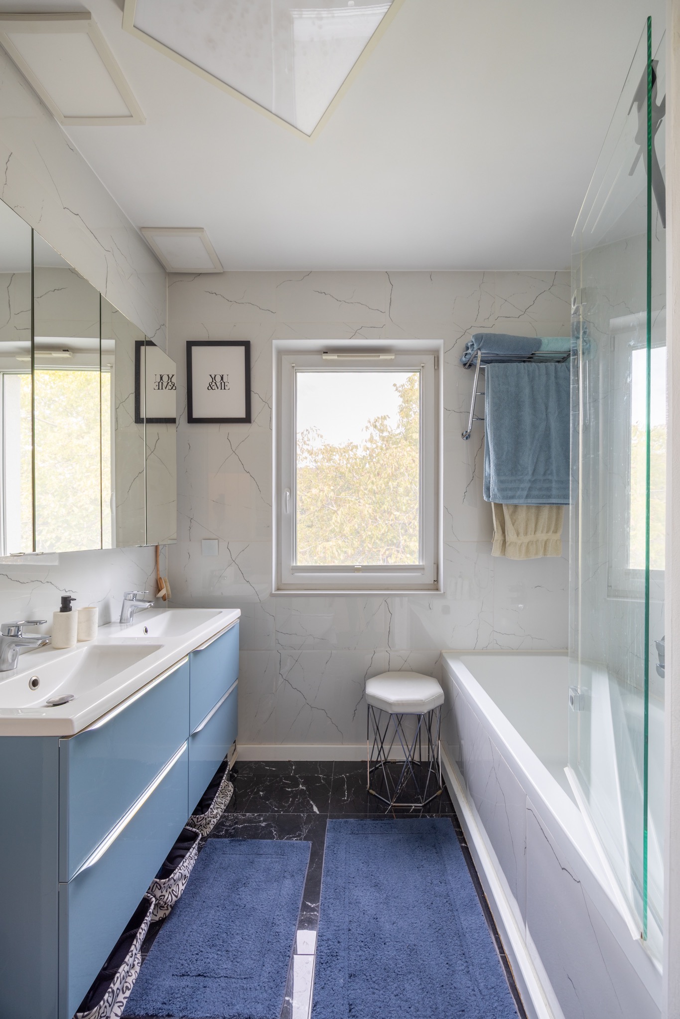 Jasna łazienka z marmurowymi płytkami, błękitną szafką pod umywalką i czarną podłogą. Okno z widokiem na zieleń. Minimalistyczny wystrój.