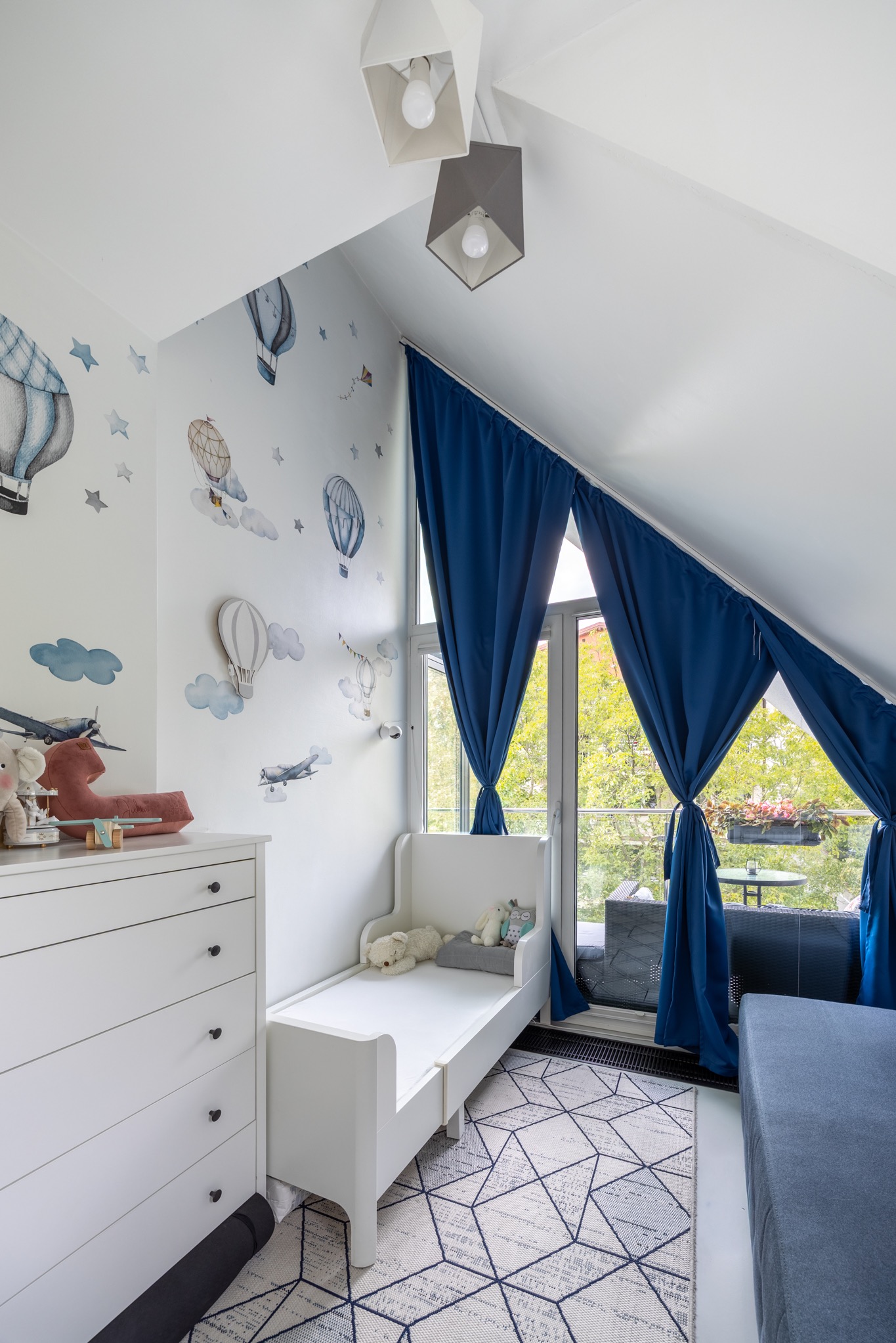 Pokój dziecięcy na poddaszu z łóżkiem, komodą i balkonem. Ściany zdobią naklejki z motywem balonów i samolotów. Widok na zieleń z balkonu.