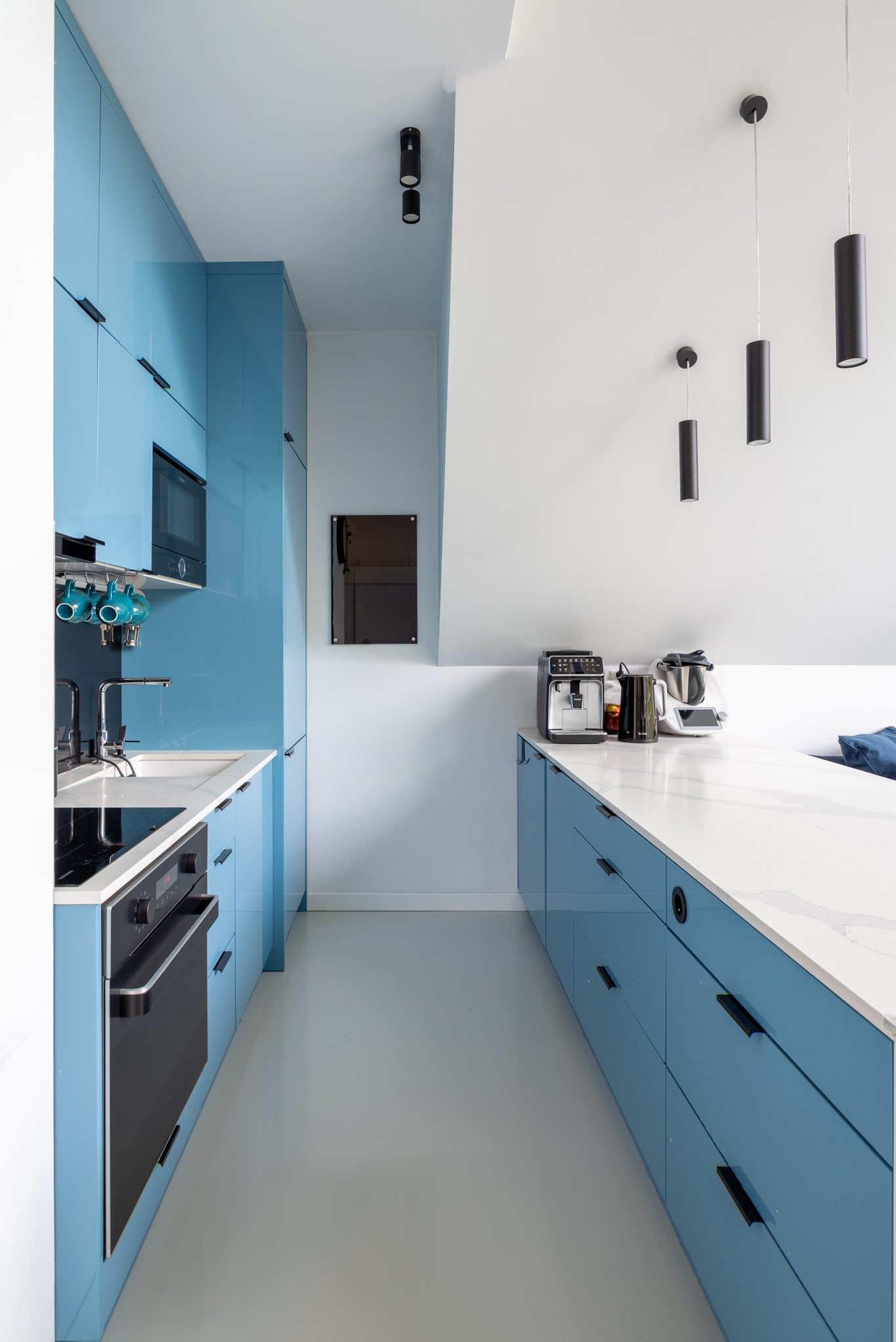 Błękitna, nowoczesna kuchnia z białym blatem i czarnymi akcentami oświetleniowymi. Widok na sprzęt AGD i minimalistyczne dodatki.