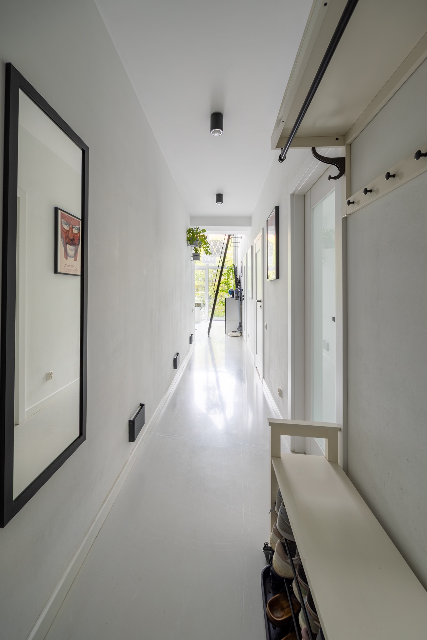 Nowoczesny, minimalistyczny korytarz w bieli z czarnymi akcentami: lustro w ramie, oświetlenie punktowe, w tle drabina i rośliny doniczkowe. Funkcjonalne i stylowe wnętrze.