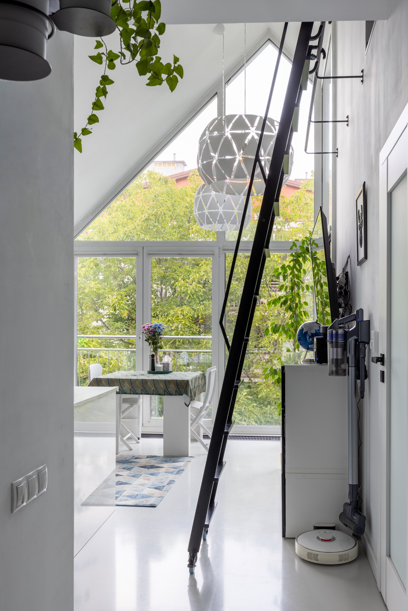 Jasne wnętrze z wysokim oknem i antresolą. W tle stół z kwiatami, a w rogu drabina i odkurzacze. Minimalistyczny wystrój z akcentami roślinnymi.