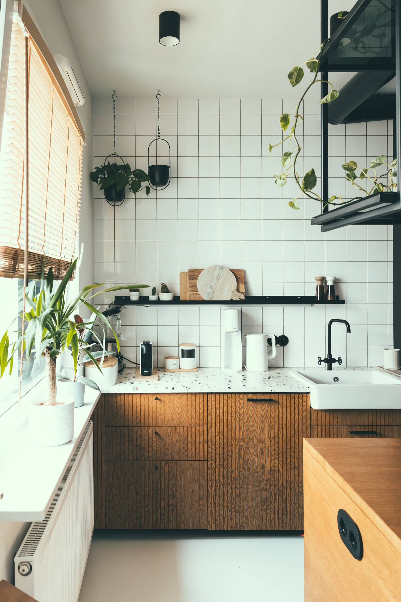 Jasna kuchnia w stylu skandynawskim z drewnianymi szafkami, białym blatem lastryko i kwadratowymi płytkami. Rośliny doniczkowe dodają świeżości.