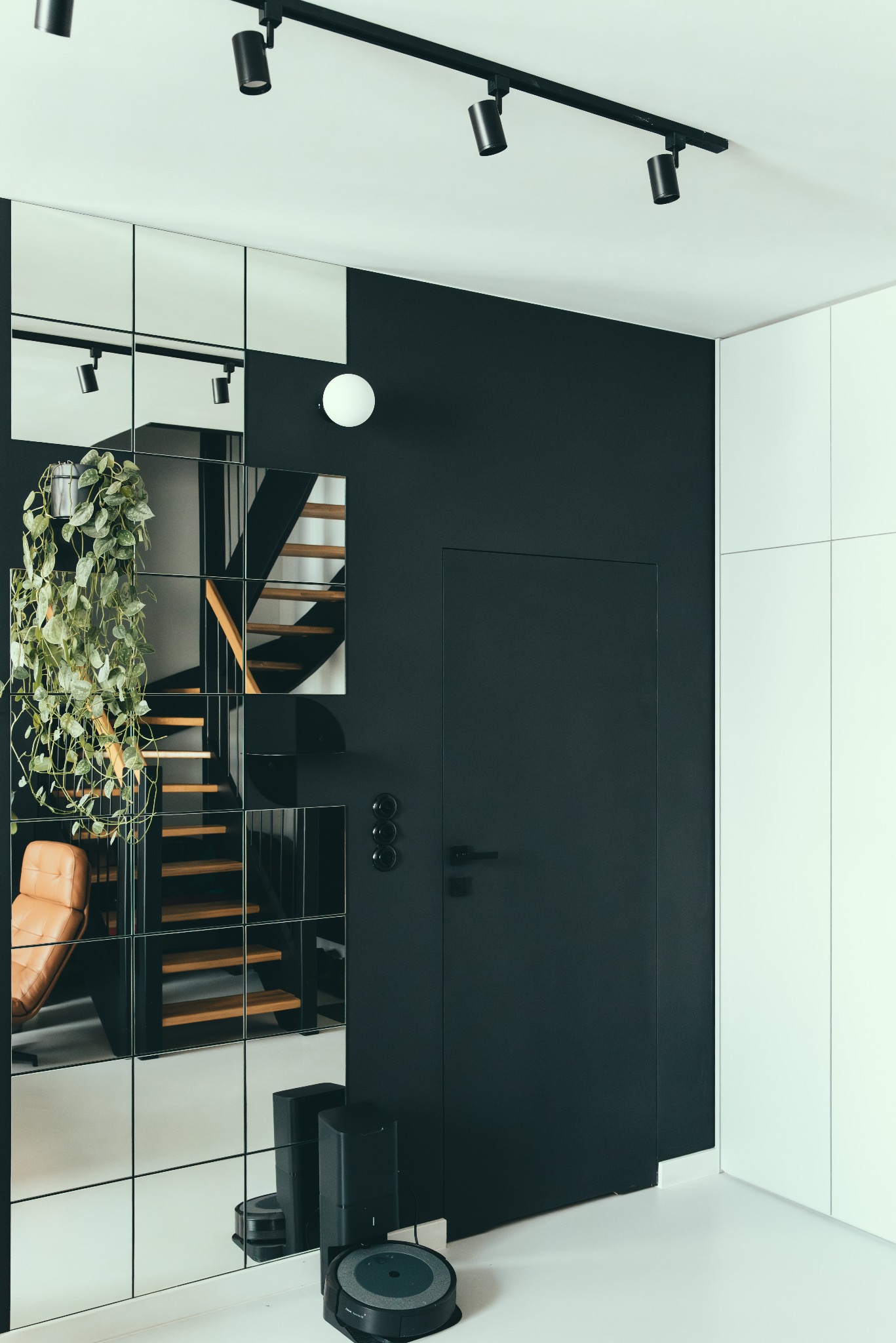 Minimalistyczne wnętrze: czarna ściana z ukrytymi drzwiami, lustrzana ściana odbijająca schody i fotel, robot sprzątający na białej podłodze.