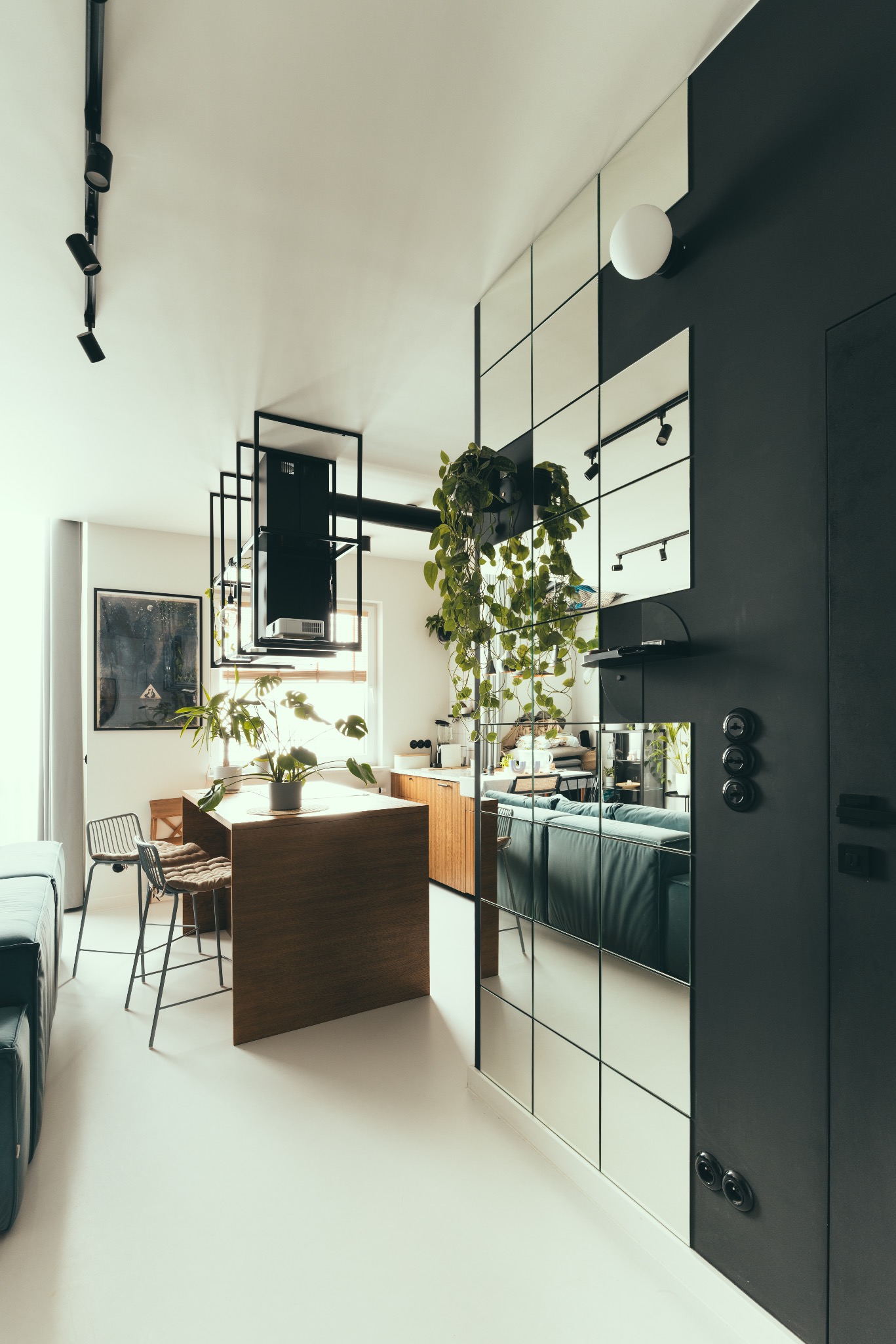 Nowoczesne wnętrze z wyspą kuchenną, roślinami i ścianą z luster. Widoczne oświetlenie punktowe i minimalistyczny design. Dominują biel, czerń i odcienie drewna.