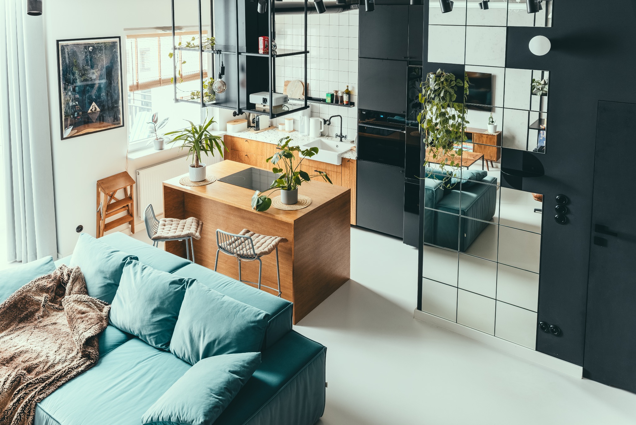 Stylowe wnętrze: turkusowa sofa z narzutą, drewniana wyspa kuchenna z hokerami i roślinami, czarne szafki, ściana z lustrami, obraz i schodki przy oknie.