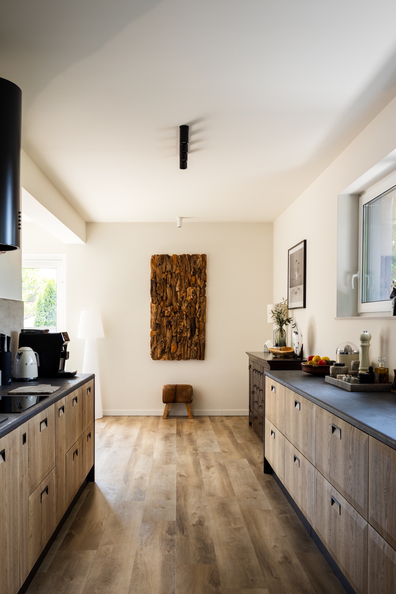 Jasna kuchnia z drewnianymi szafkami, czarnym blatem i elementami dekoracyjnymi: lampa, obraz z drewna, stołek. Wnętrze doświetlone naturalnym światłem.