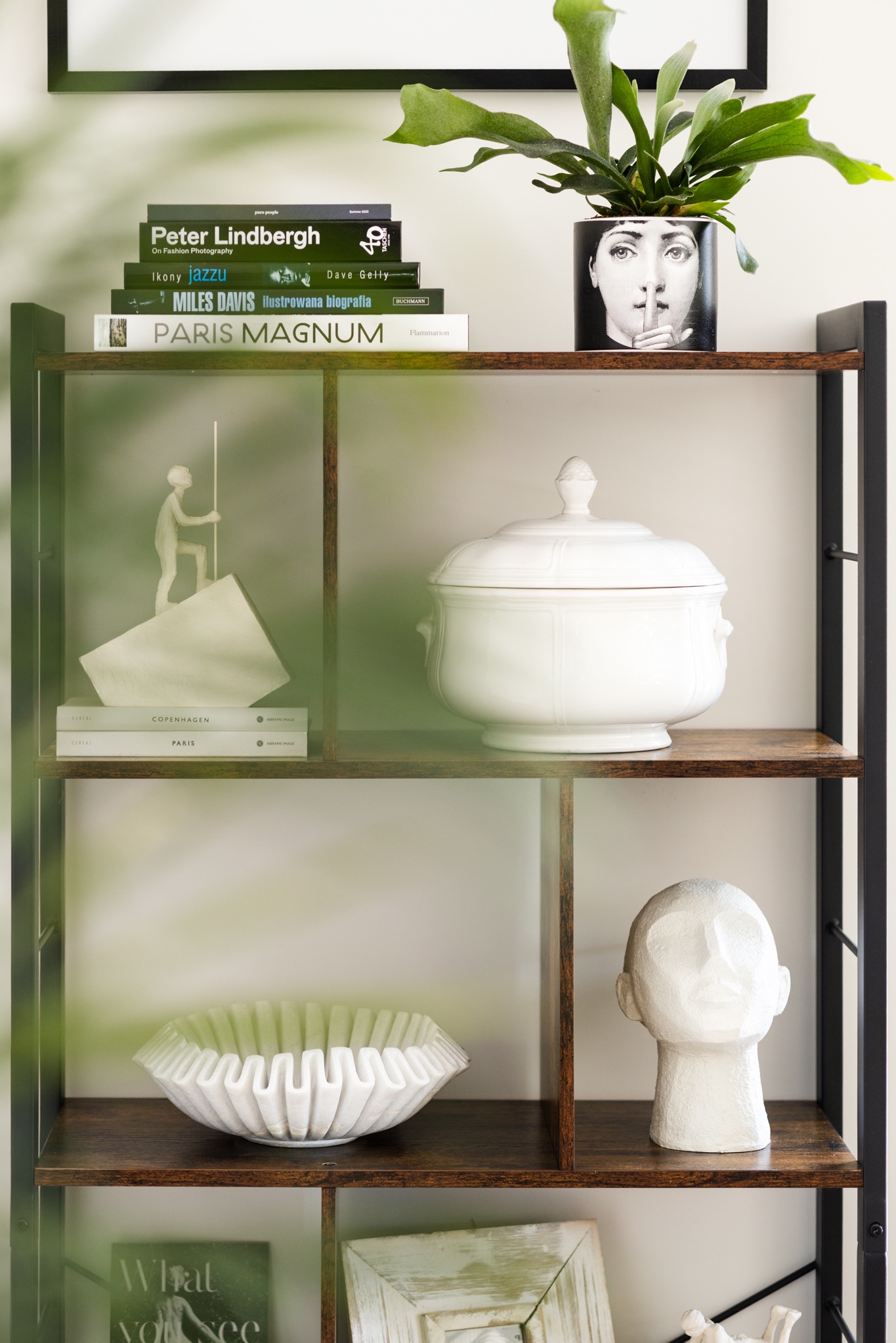 Regał z dekoracjami: książki (Lindbergh, Davis, Paris Magnum), rzeźby (głowa, figurka), misa i doniczka z motywem twarzy. Nowoczesny design wnętrza.