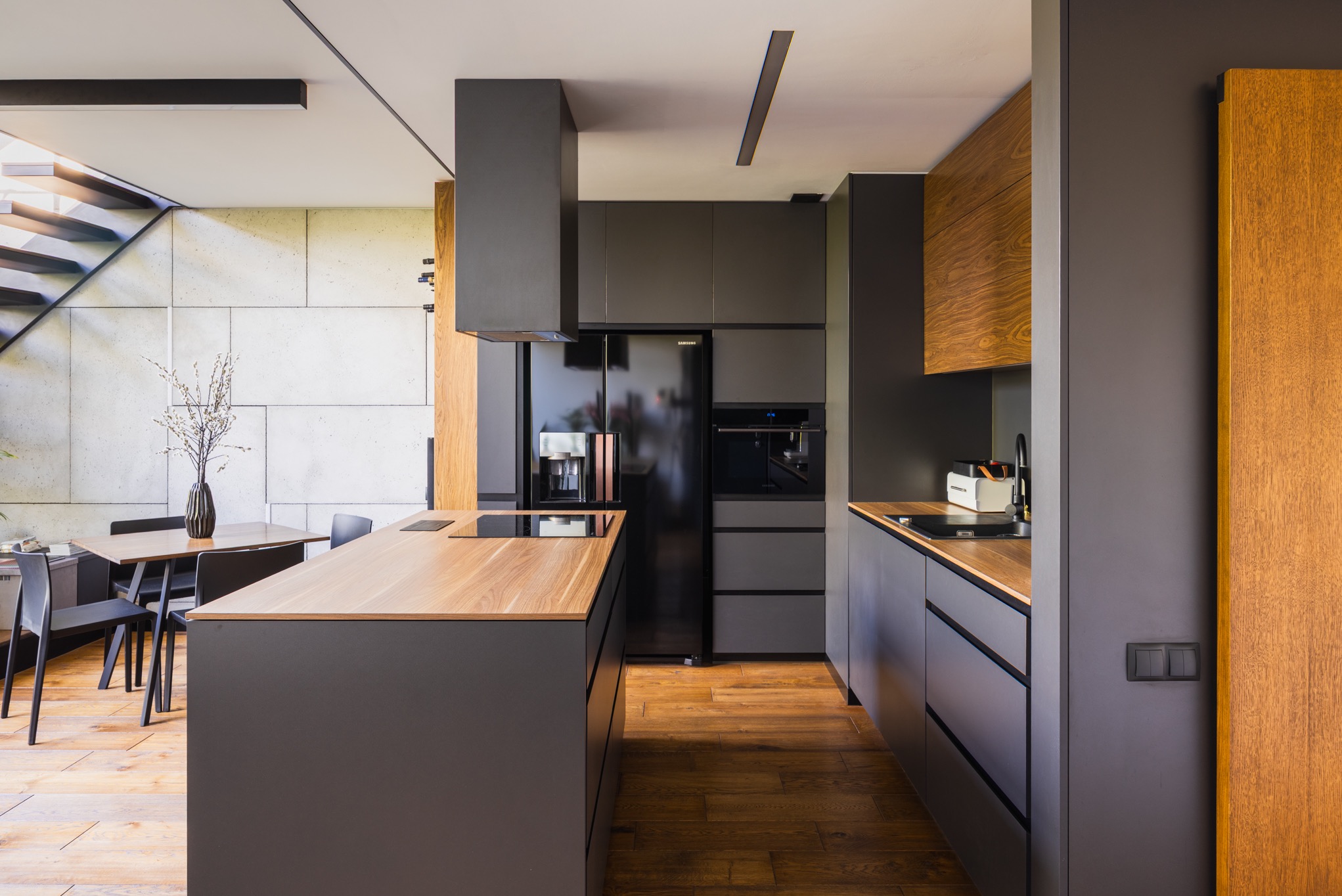 Nowoczesna, szaro-drewniana kuchnia z wyspą i schodami w tle, uchwycona szerokim kątem w Warszawie. Minimalistyczny design, drewniane blaty i podłoga.