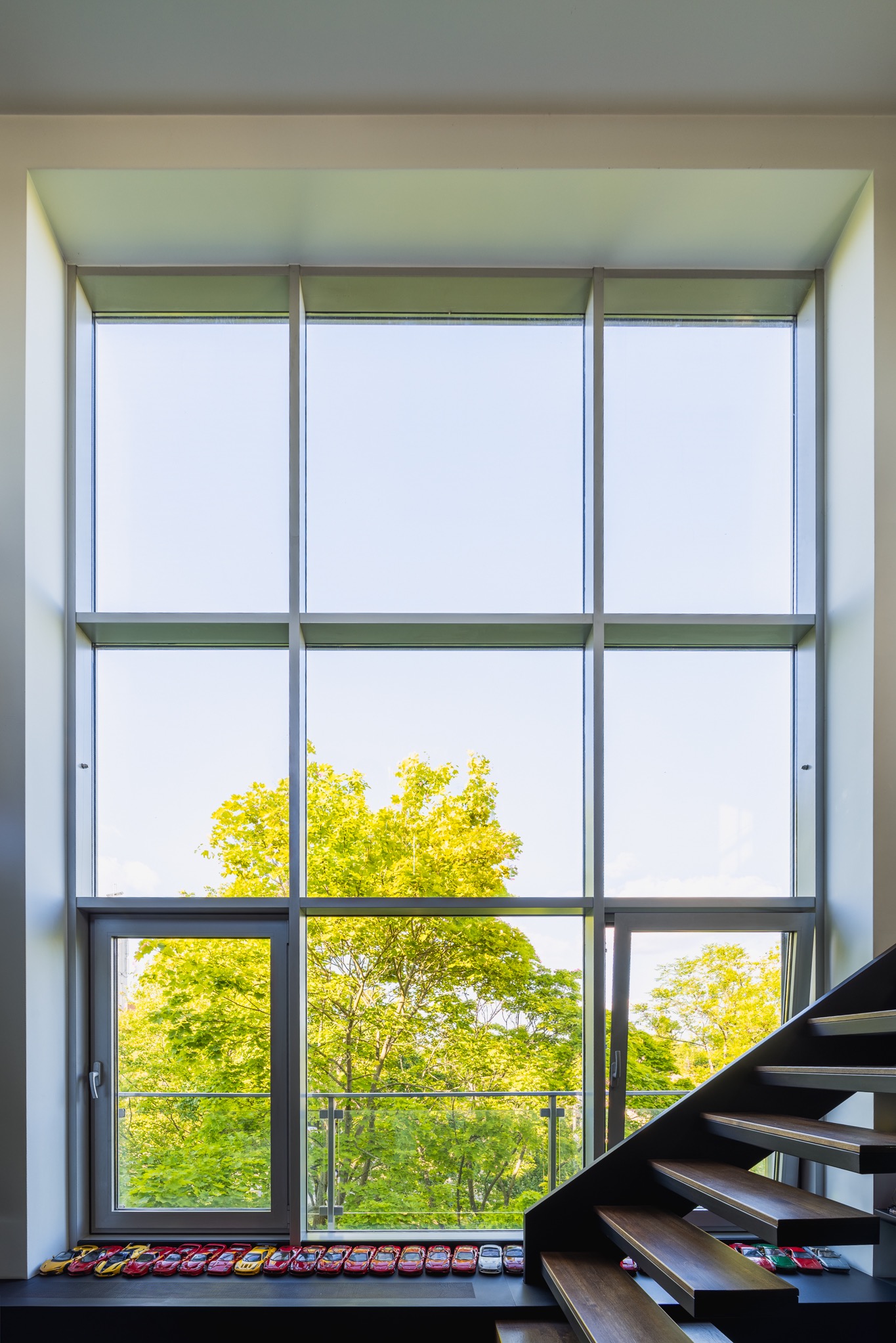 Wysokie okno z widokiem na zieleń, obok schody z drewnianymi stopniami. Na parapecie kolekcja modeli samochodów. Jasne, naturalne światło.