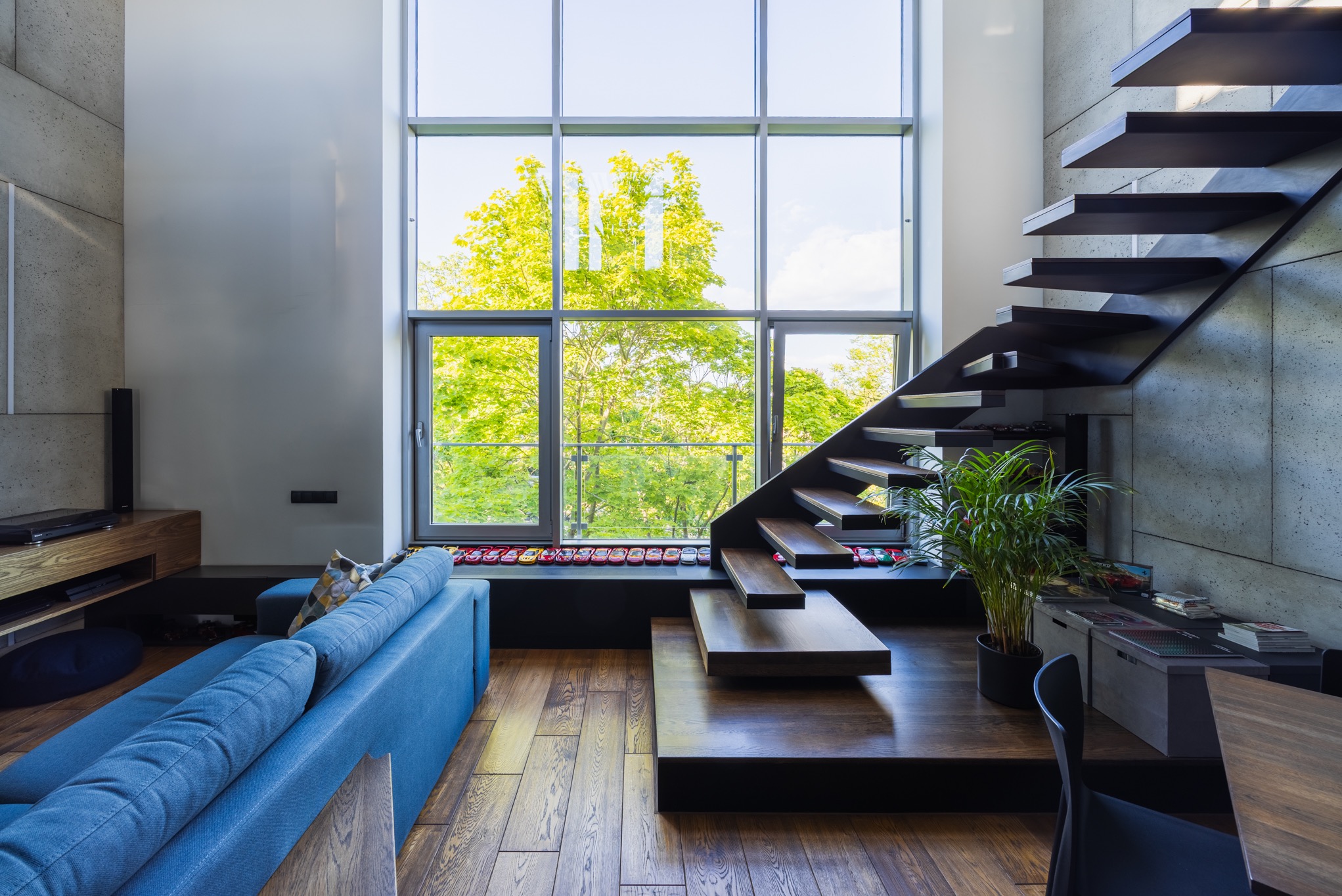Nowoczesne wnętrze z otwartymi schodami, dużym oknem z widokiem na zieleń i niebieską sofą. Na parapecie kolekcja modeli samochodów.