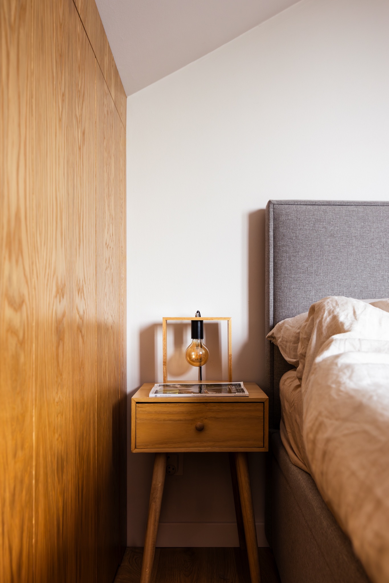 Sypialnia z drewnianą szafką nocną, lampką z żarówką Edisona i magazynem. Minimalistyczny wystrój wnętrza z szarym zagłówkiem łóżka.