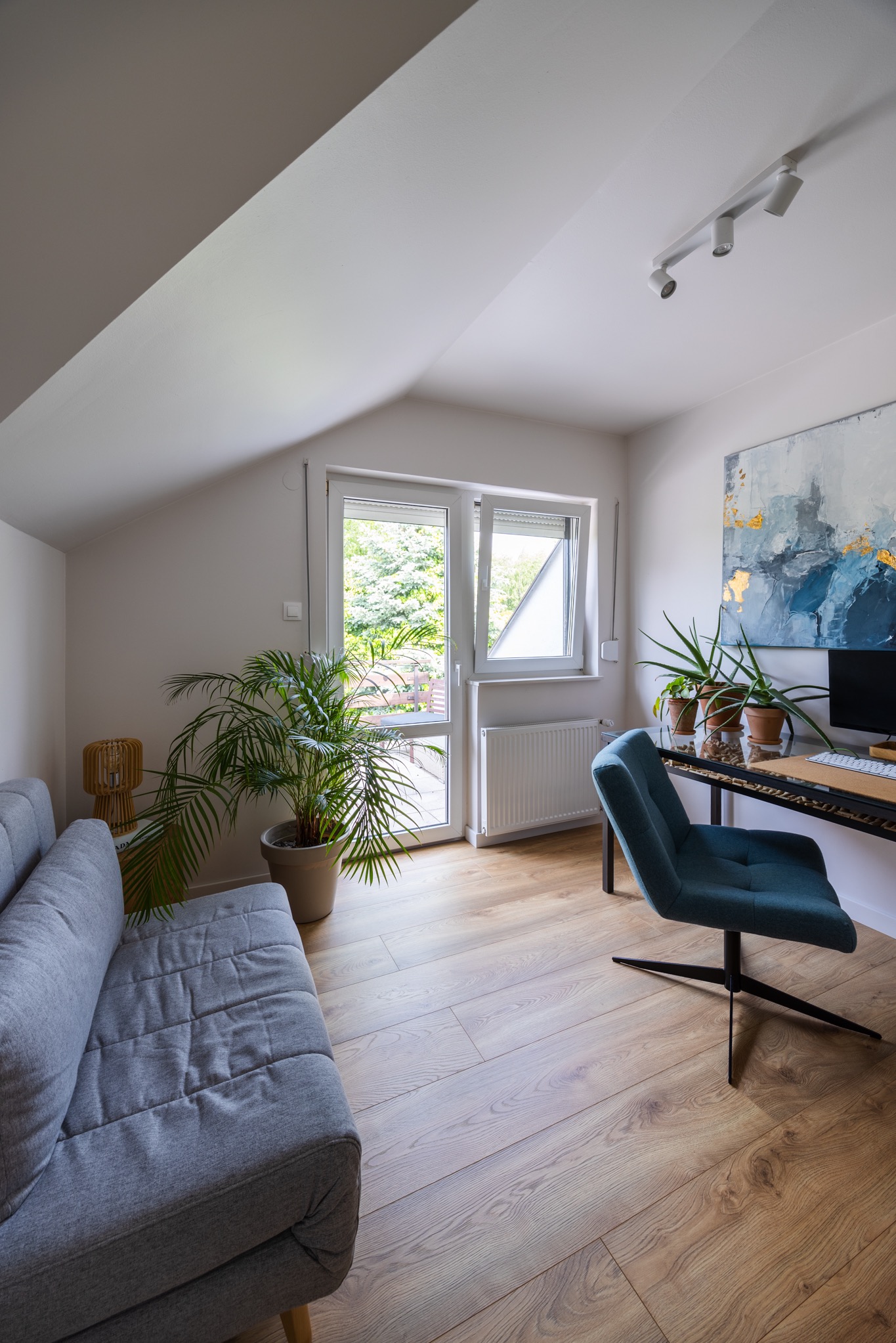 Jasne poddasze: szara sofa, biurko z komputerem, rośliny doniczkowe i otwarte okno z widokiem na zieleń. Abstrakcyjny obraz z akcentami złota na ścianie.