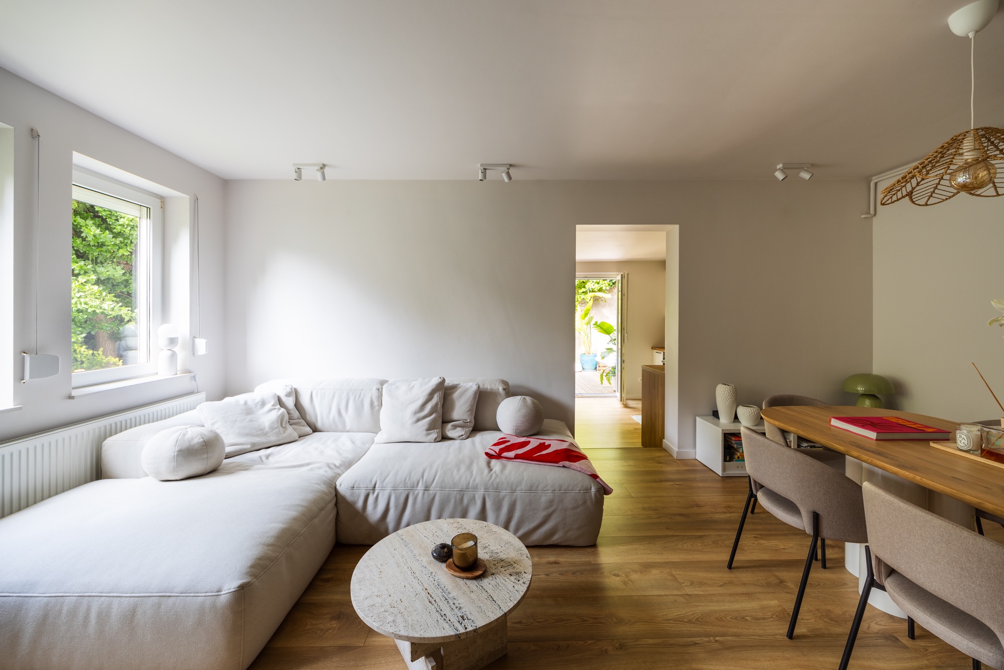 Jasne wnętrze: narożnik z poduszkami, okrągły stolik z trawertynu, drewniany stół z krzesłami. Widok na ogród przez otwarte drzwi. Minimalistyczny design.