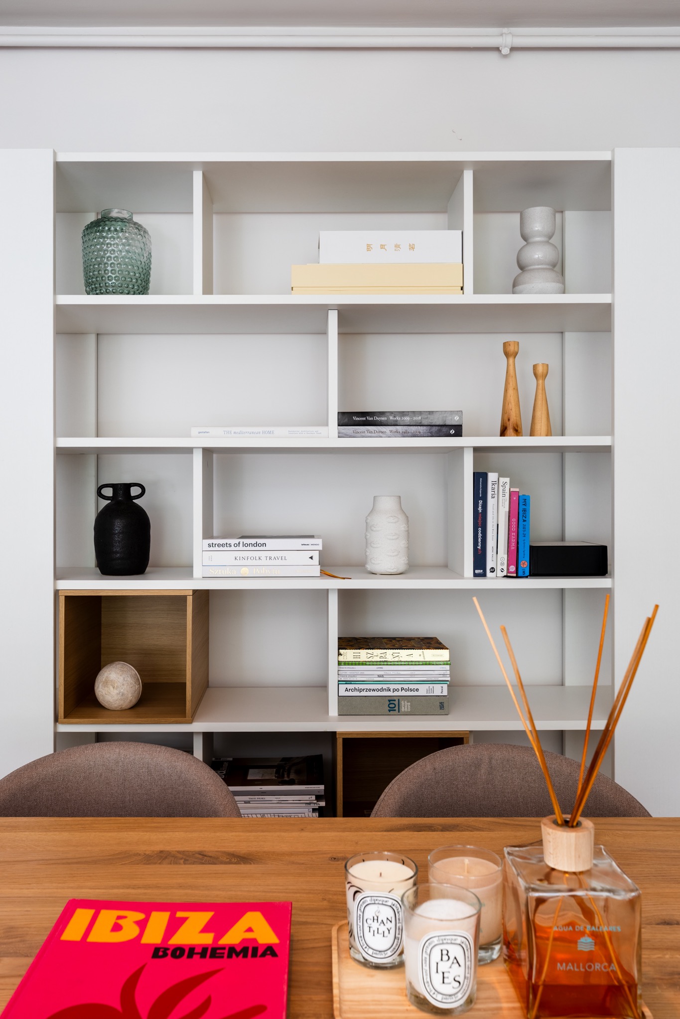Biały regał z dekoracjami: wazony, książki i świece na drewnianym stole. Minimalistyczny wystrój wnętrza z akcentami kolorystycznymi i dyfuzorem zapachowym.