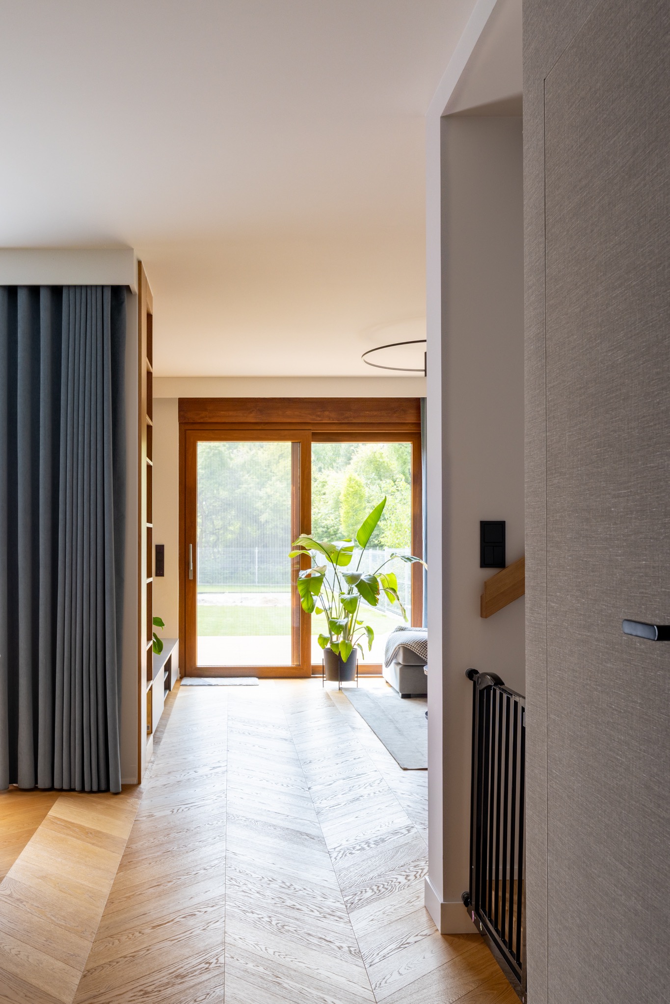 Jasne wnętrze domu z parkietem w jodełkę, drewnianymi drzwiami balkonowymi i rośliną doniczkową. Widok z korytarza, brama zabezpieczająca na schodach.