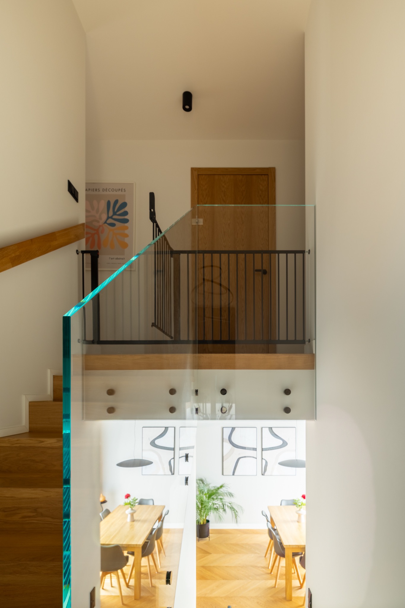 Nowoczesne wnętrze domu z drewnianymi schodami i szklaną balustradą z turkusowym akcentem, widok z góry na jadalnię z parkietem w jodełkę.