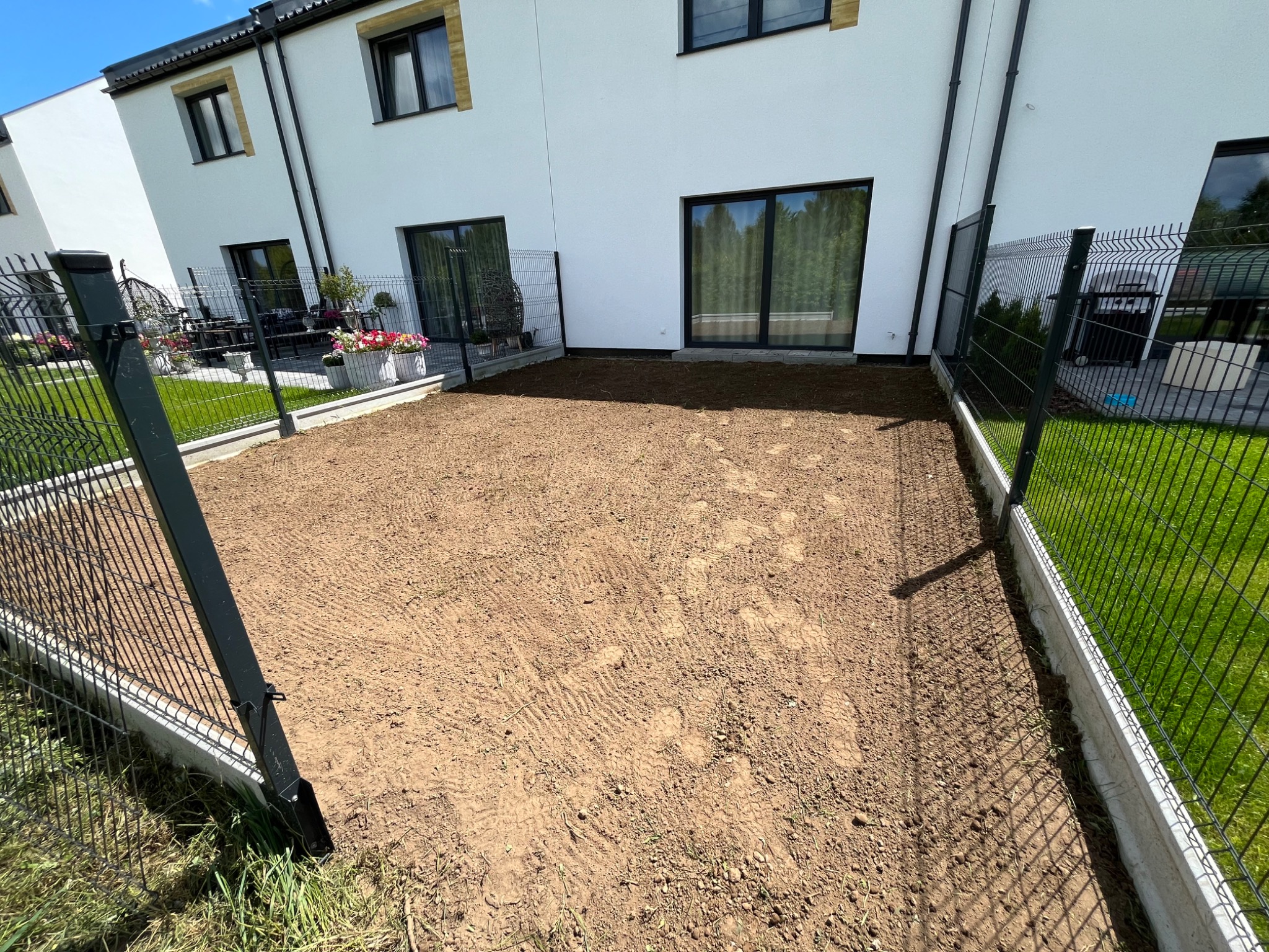 Przygotowany teren pod trawnik z widocznymi śladami po maszynach, ogrodzony siatką na betonowej podmurówce. W tle białe budynki szeregowe.