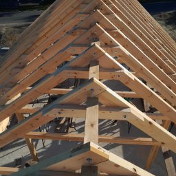 Usługi Dekarskie Piotr Zaremba - Drewniana więźba dachowa w trakcie budowy, widok z góry, z widocznymi elementami konstrukcyjnymi i śrubami mocującymi. W tle budynek mieszkalny.