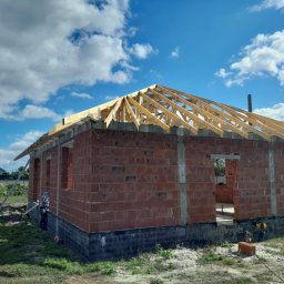 Usługi Dekarskie Piotr Zaremba - Wznoszenie więźby dachowej na murowanym budynku w stanie surowym otwartym, z widoczną konstrukcją drewnianą i fragmentem nieba z chmurami. Prace budowlane w toku.