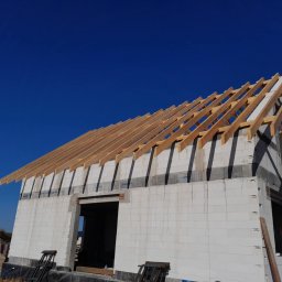 Usługi Dekarskie Piotr Zaremba - Budowa domu z białych bloczków, widoczna drewniana więźba dachowa na tle intensywnego, błękitnego nieba. Ujęcie z dołu, podkreślające konstrukcję dachu.