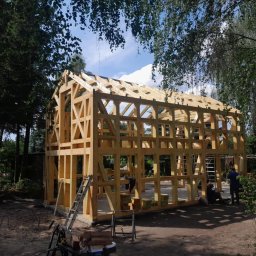 Usługi Dekarskie Piotr Zaremba - Konstrukcja drewniana domu w trakcie budowy, widoczne elementy więźby dachowej na tle zieleni drzew i błękitnego nieba. Na ziemi leżą materiały budowlane.