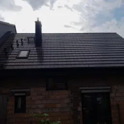 Dom z ciemną, nową dachówką ceramiczną, widoczne okno dachowe i komin z ozdobnym zwieńczeniem na tle pochmurnego nieba.