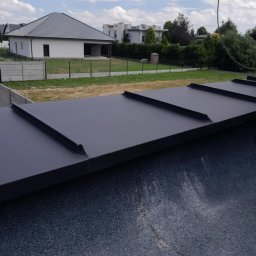 Usługi Dekarskie Piotr Zaremba - Szczegółowe ujęcie fragmentu dachu z blachy na tle zabudowań i błękitnego nieba. Widoczne łączenia blachy, faktura materiału i elementy wykończeniowe.