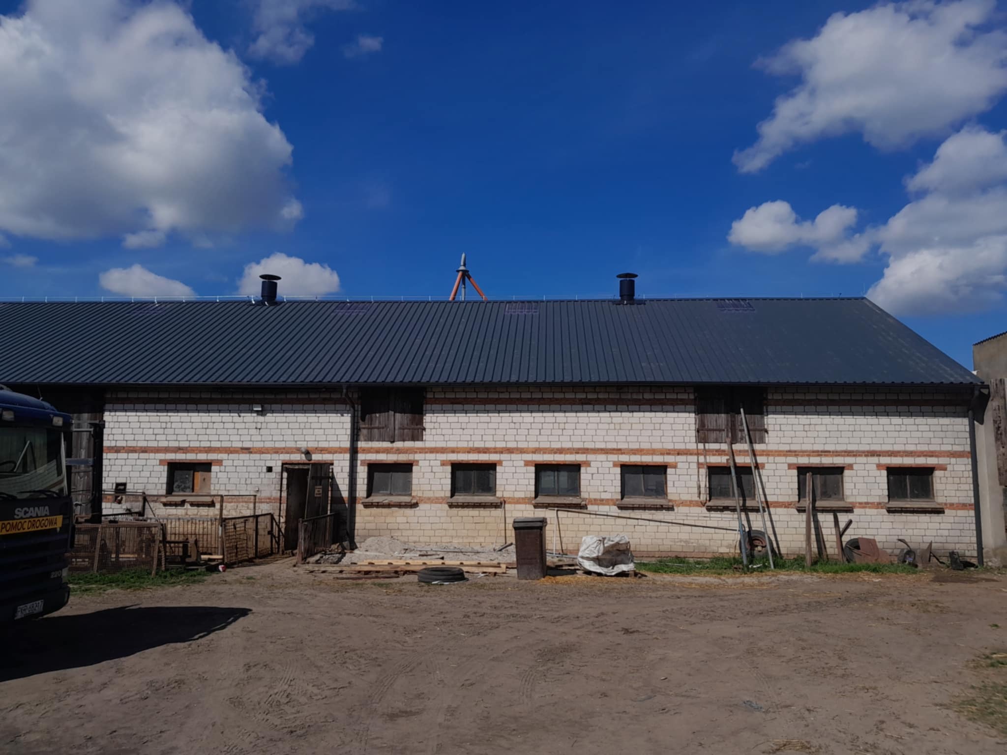 Długi, murowany budynek gospodarczy z nowym, ciemnym dachem z blachy trapezowej. Na dachu widoczne kominy i maszt. Błękitne niebo z białymi obłokami w tle.