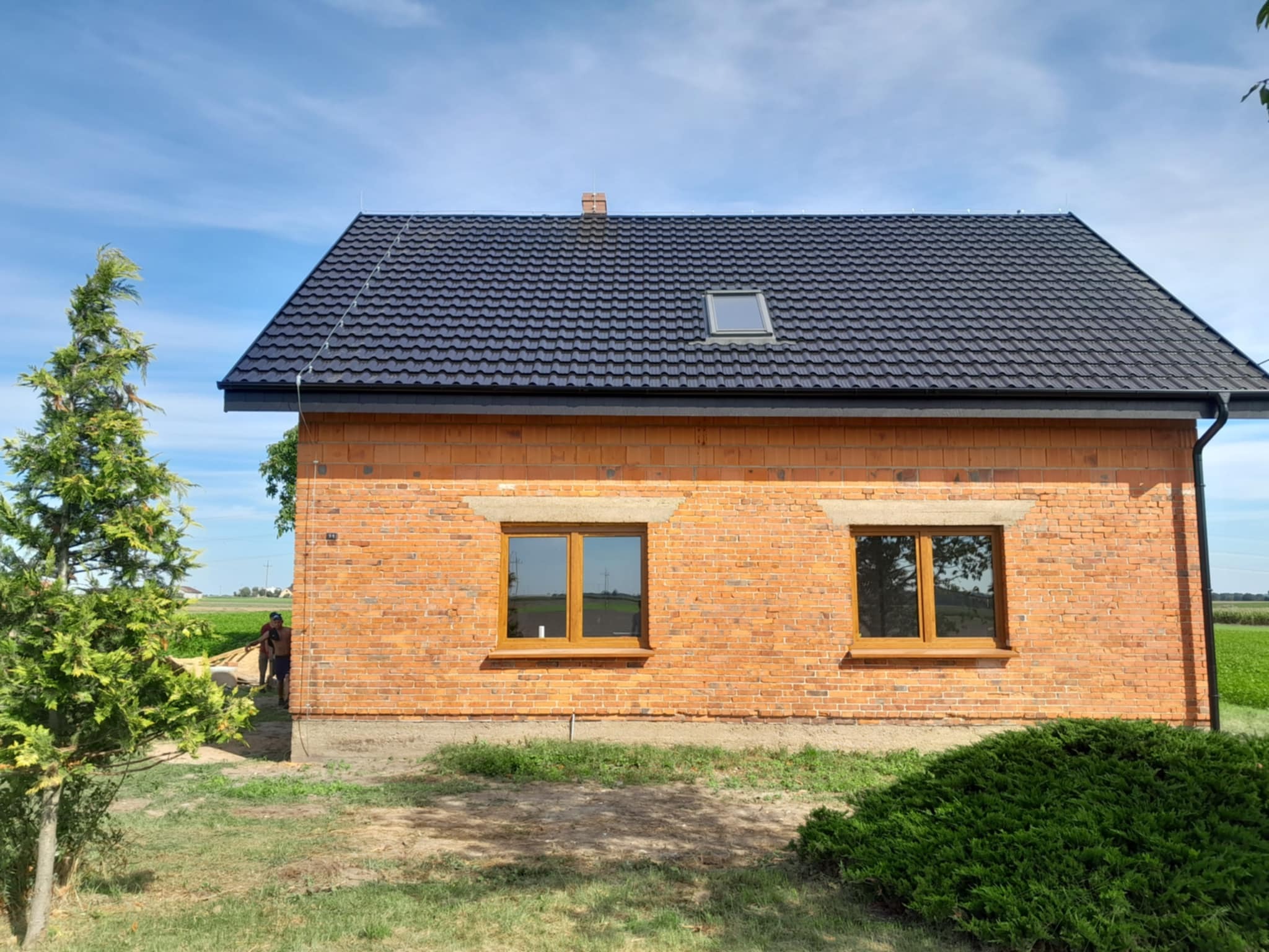 Niedokończony dom z cegły z nowym, czarnym dachem z dachówki i oknem dachowym. Widoczna rynna, fragment elewacji i osoby w tle. Budynek w otoczeniu zieleni.