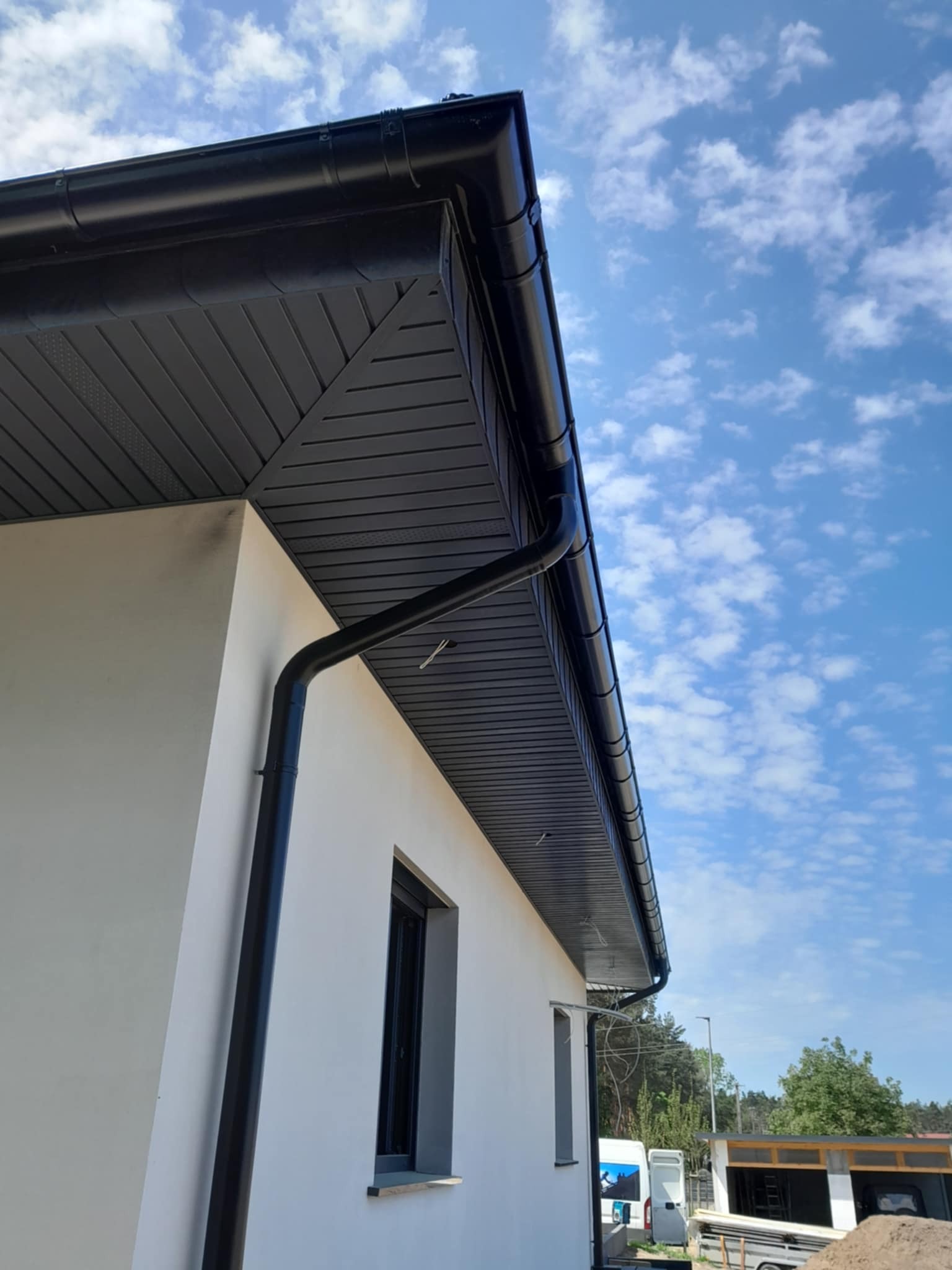 Nowoczesny dom z czarnymi rynnami i podbitką dachową na tle błękitnego nieba z chmurami. Ujęcie z dołu podkreśla geometrię budynku i kontrast kolorów.