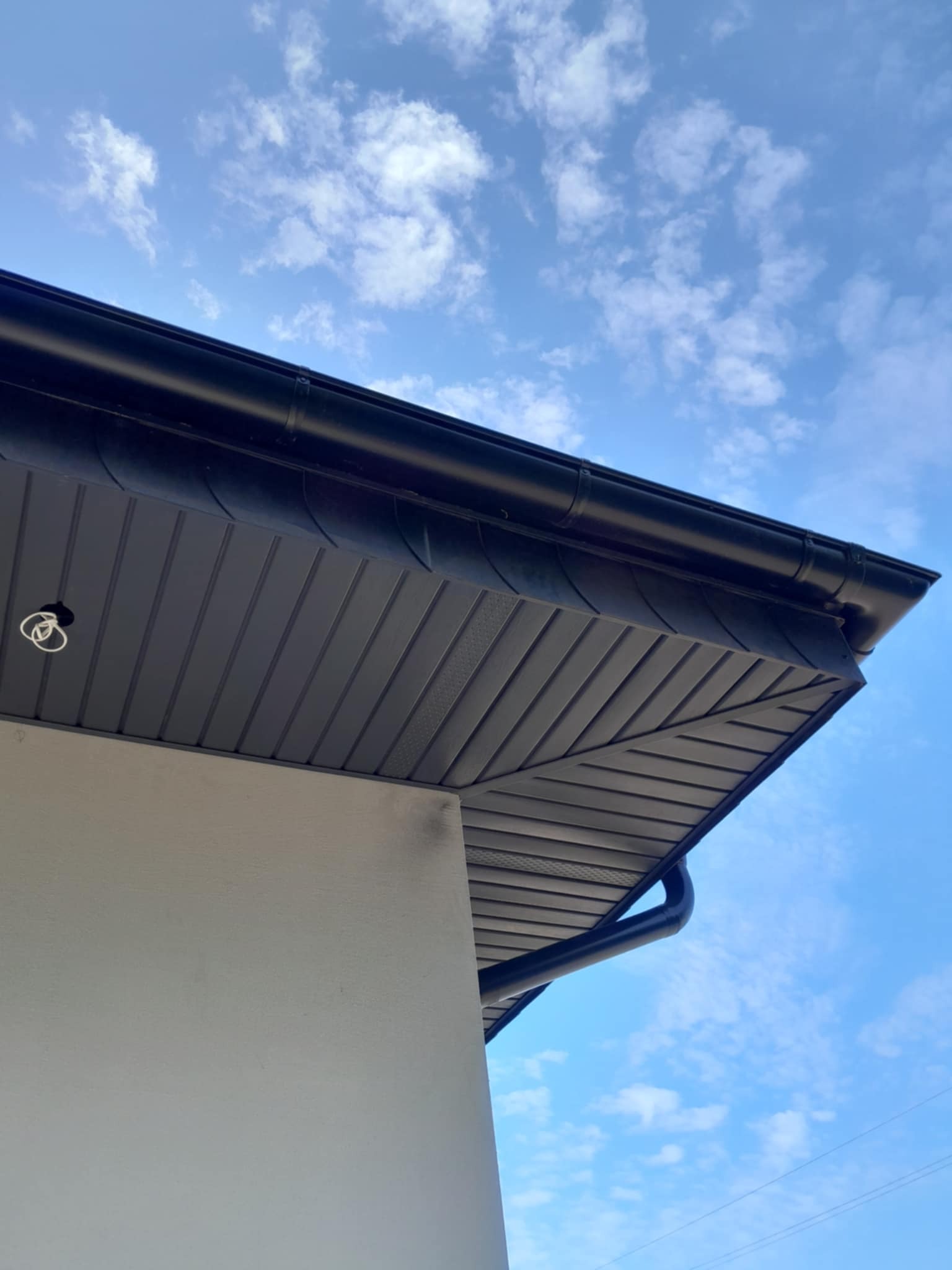 Czarne, metalowe rynny na tle błękitnego nieba z chmurami. Widoczny fragment elewacji budynku z podbitką dachową. Ujęcie z dołu, podkreślające detale.