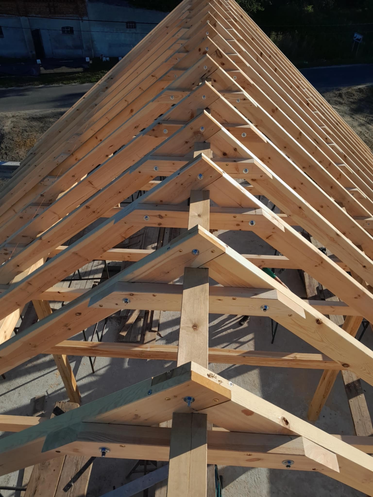 Drewniana więźba dachowa w trakcie budowy, widok z góry, z widocznymi elementami konstrukcyjnymi i śrubami mocującymi. W tle budynek mieszkalny.