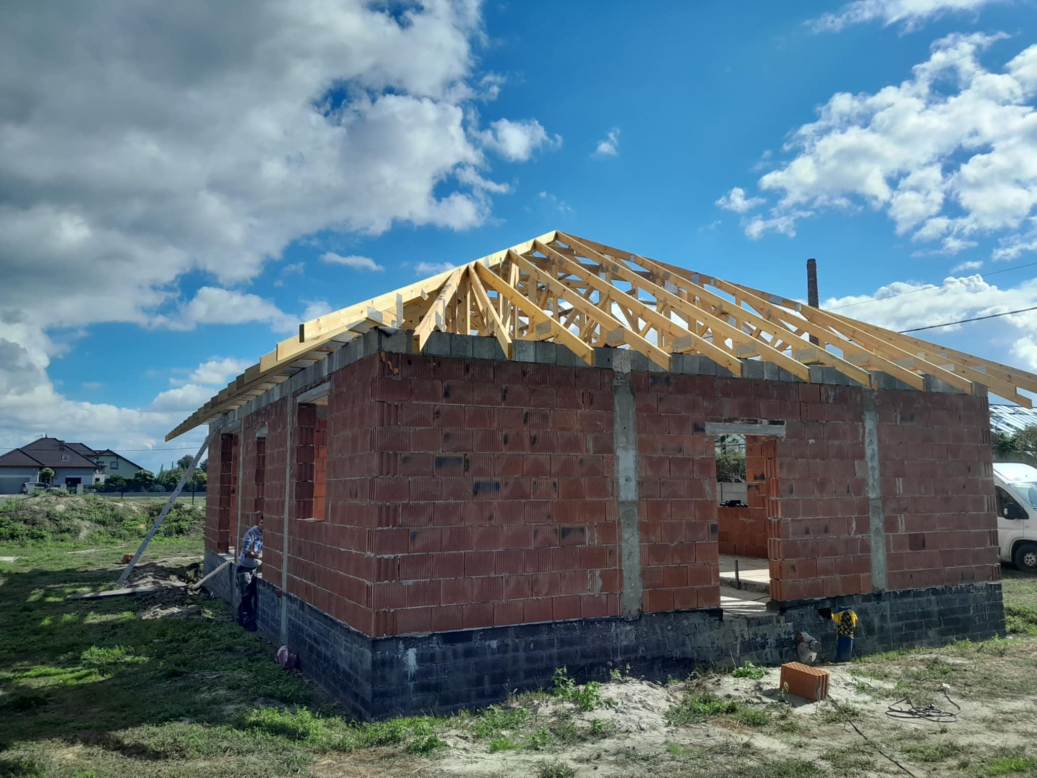 Wznoszenie więźby dachowej na murowanym budynku w stanie surowym otwartym, z widoczną konstrukcją drewnianą i fragmentem nieba z chmurami. Prace budowlane w toku.