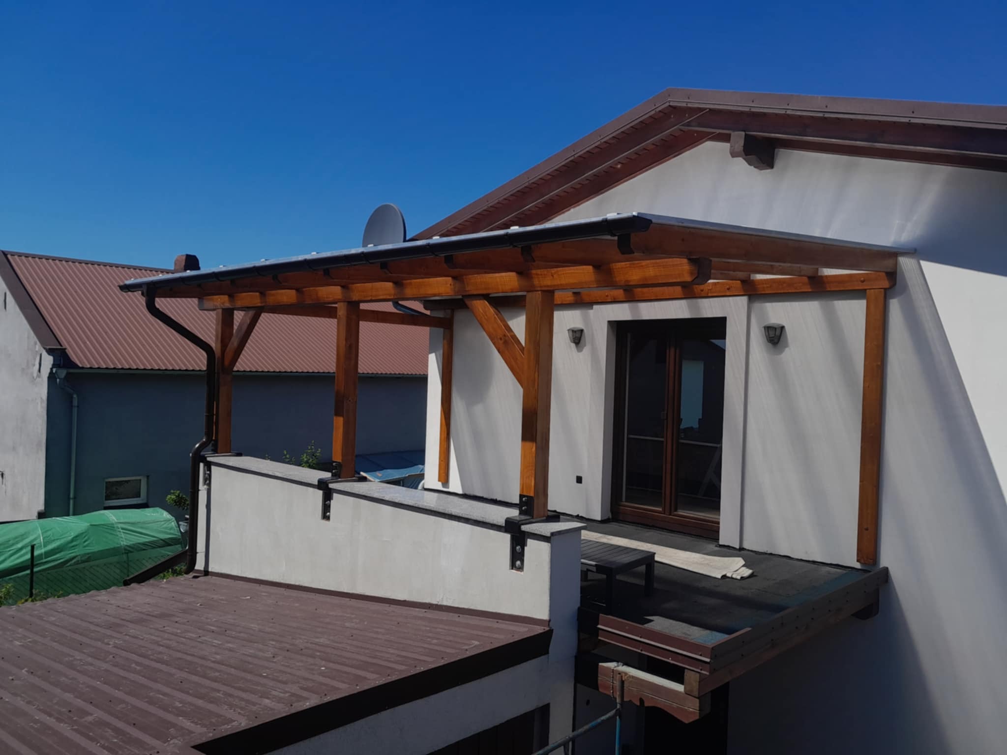 Drewniana konstrukcja zadaszenia tarasu przy budynku z brązowym dachem i rynnami. Widoczne elementy mocujące i fragment dachu sąsiedniego budynku.