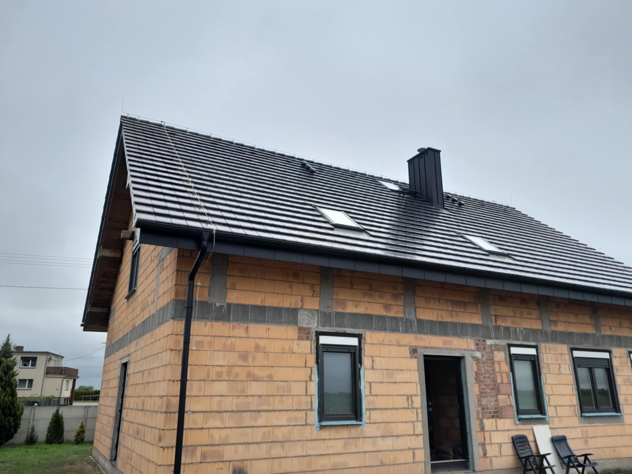 Dom w trakcie budowy w Jasnym Polu: dach z ciemnej dachówki ceramicznej z oknami dachowymi i czarną rynną, ściany z cegły. Widok z zewnątrz.