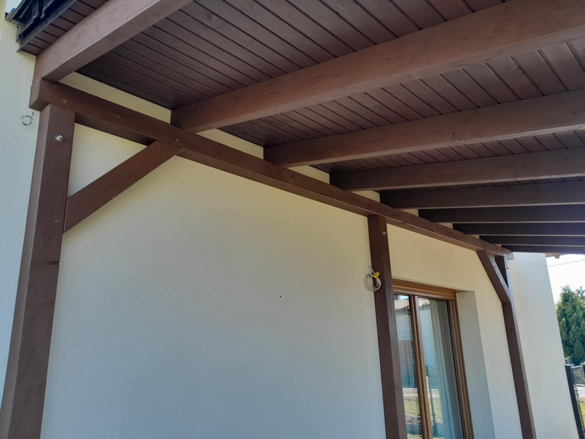 Drewniana konstrukcja z belek, prawdopodobnie zadaszenie tarasu, pomalowana na brązowo, z widocznymi elementami mocującymi i fragmentem okna w tle.