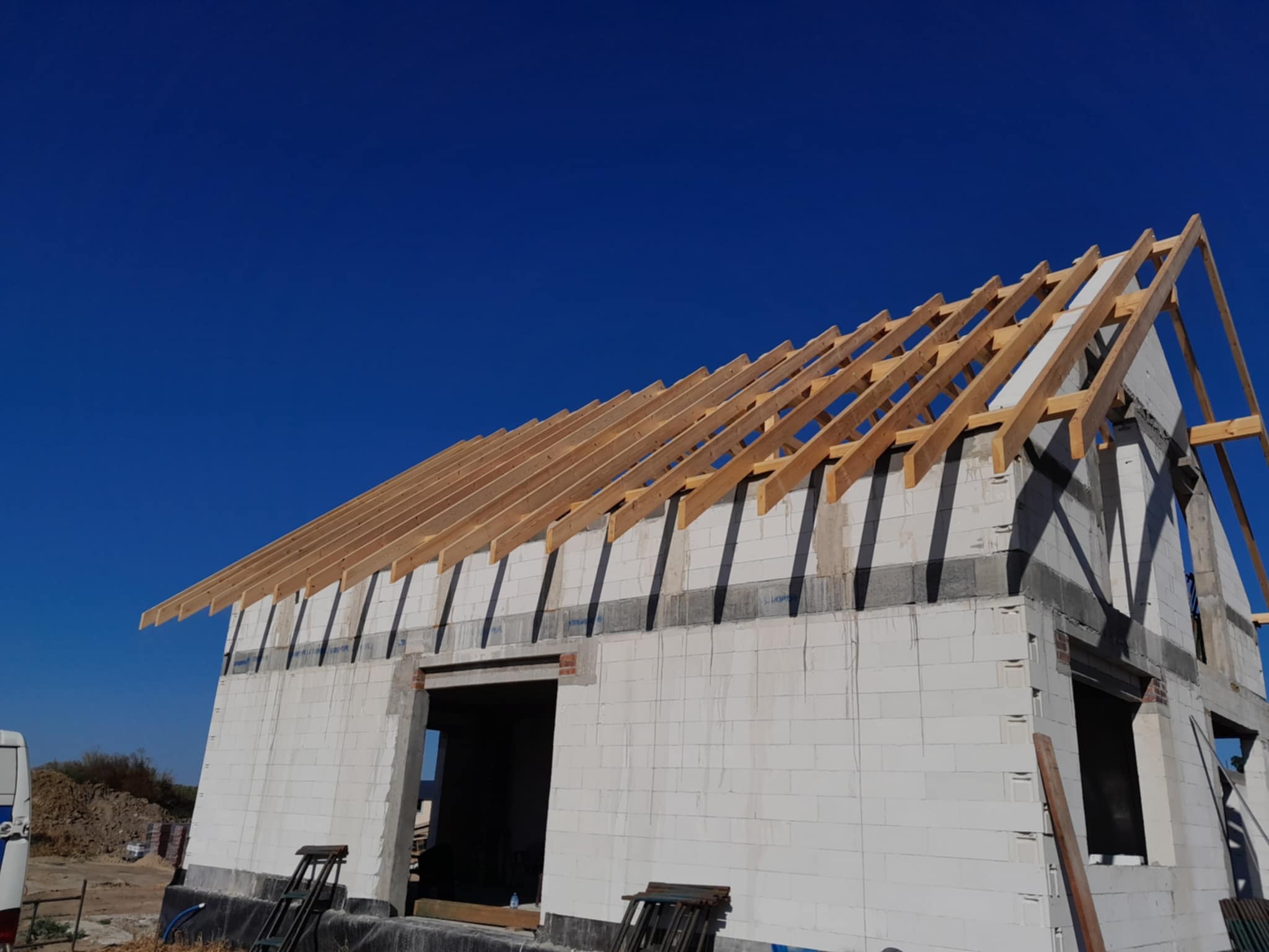 Budowa domu z białych bloczków, widoczna drewniana więźba dachowa na tle intensywnego, błękitnego nieba. Ujęcie z dołu, podkreślające konstrukcję dachu.