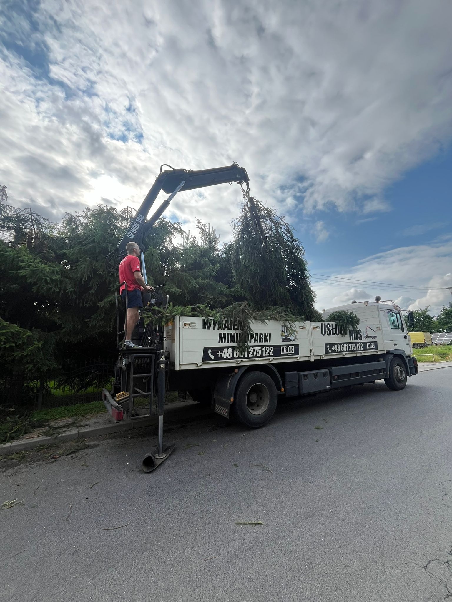 Usługi HDS: Wywóz gałęzi z przyczepy za pomocą dźwigu HDS, operator w czerwonej koszulce, błękitne niebo z chmurami w tle.