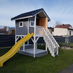 Mateusz Stronczek - Drewniany domek dla dzieci z żółtą zjeżdżalnią i białymi schodami na trawniku. Szary domek z ciemnym dachem i małymi oknami. Tło stanowią budynki mieszkalne.
