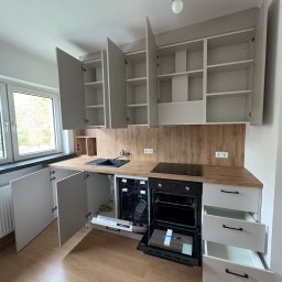 Mateusz Stronczek - Jasna kuchnia z drewnianym blatem i otwartymi szafkami, ukazująca wnętrze i sprzęt AGD, w tym piekarnik i zmywarkę. Widoczne detale wykończenia i oświetlenie.