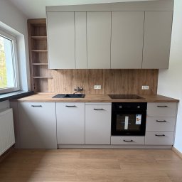 Mateusz Stronczek - Jasna kuchnia z drewnianym blatem i panelami. Widoczne szafki wiszące i stojące, piekarnik, płyta indukcyjna, zlew i okno. Podłoga wyłożona panelami imitującymi drewno.