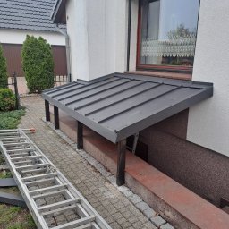 Mateusz Stronczek - Zadaszenie nad zejściem do piwnicy z szarej blachy trapezowej, wsparte na drewnianych słupkach. Aluminiowa drabina oparta o chodnik z kostki brukowej.