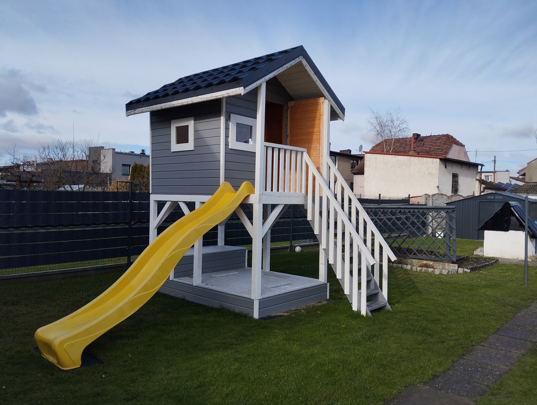 Drewniany domek dla dzieci z żółtą zjeżdżalnią i białymi schodami na trawniku. Szary domek z ciemnym dachem i małymi oknami. Tło stanowią budynki mieszkalne.