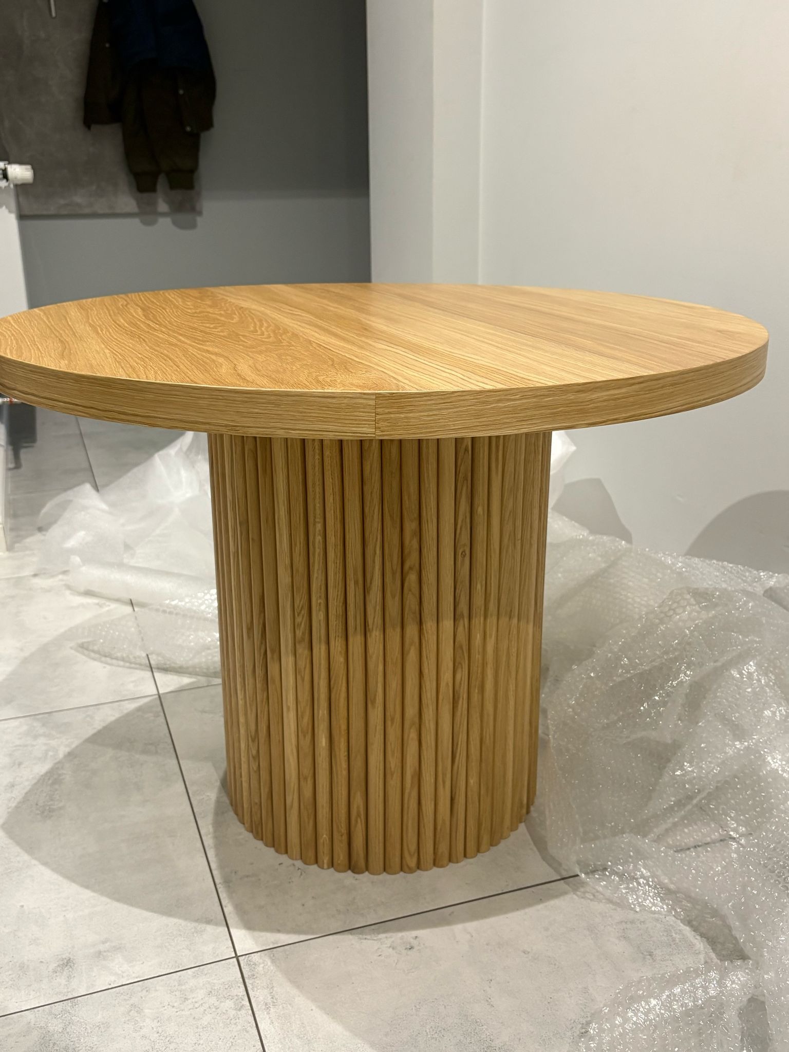 Okrągły, drewniany stół z żłobioną podstawą na tle jasnych płytek i folii bąbelkowej. Minimalistyczny design, ciepłe odcienie drewna, nowoczesny styl.