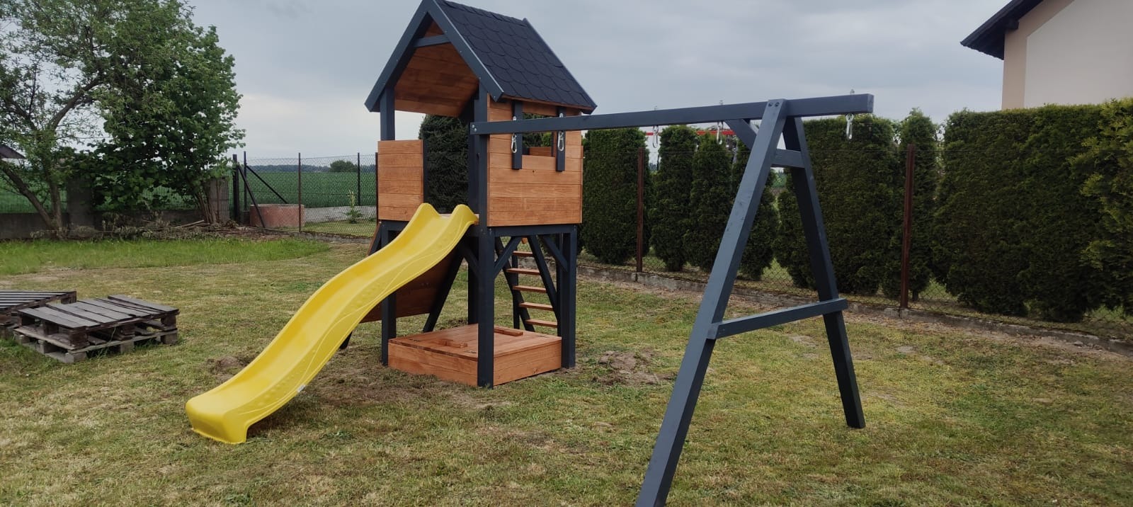 Drewniany plac zabaw z wieżą, zjeżdżalnią i huśtawką, pomalowany na szaro i brązowo, na tle zielonego trawnika i żywopłotu. Widoczne palety obok konstrukcji.