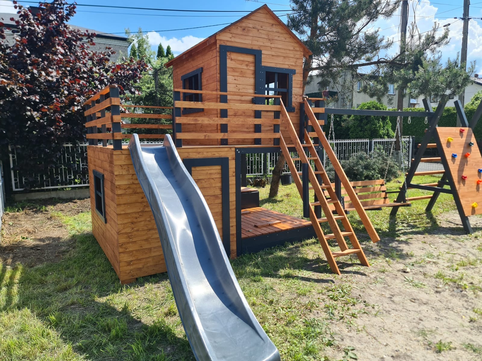 Drewniany plac zabaw z domkiem, zjeżdżalnią, drabinką i ścianką wspinaczkową na trawniku w ogrodzie, w słoneczny dzień. Elementy pomalowane na brązowo i czarno.