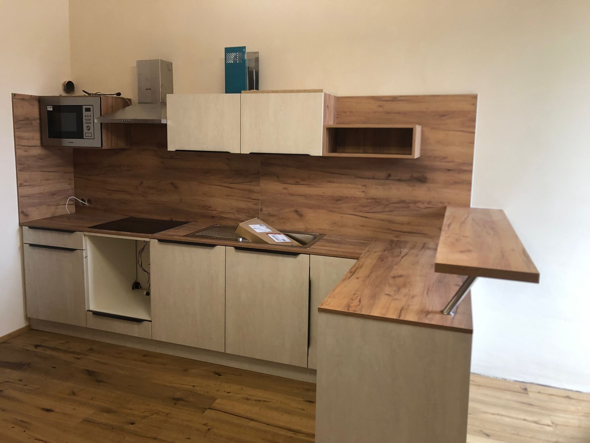 Jasna kuchnia z drewnianymi akcentami: zabudowa z mikrofalówką, okapem i barkiem. Widoczne niedokończone elementy, brakujące fronty szafek i karton w zlewie.