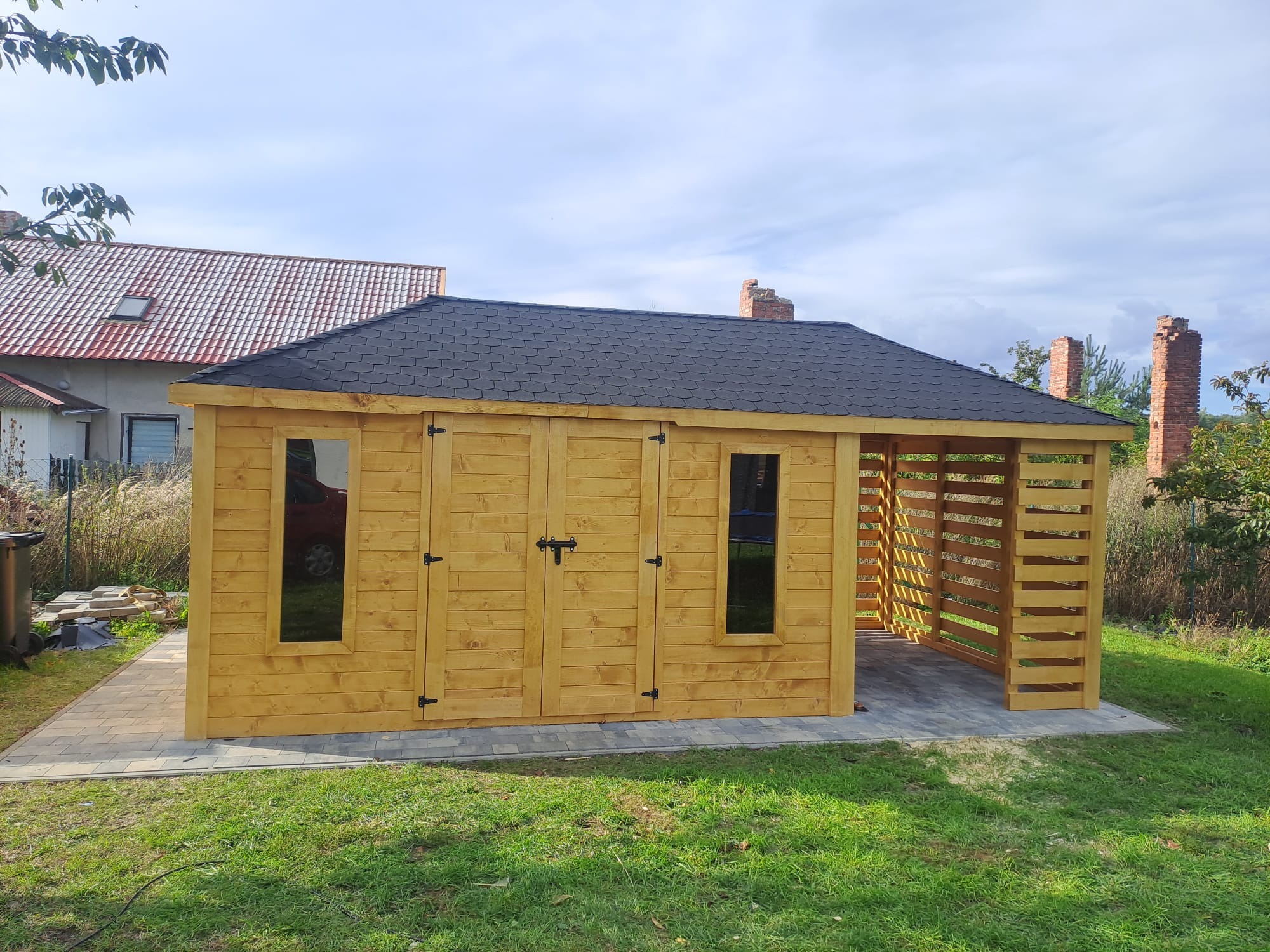 Drewniany domek narzędziowy z dwuspadowym dachem krytym gontem bitumicznym, z oknami i podwójnymi drzwiami, zadaszony taras boczny, na kostce brukowej, otoczony zielonym trawnikiem.