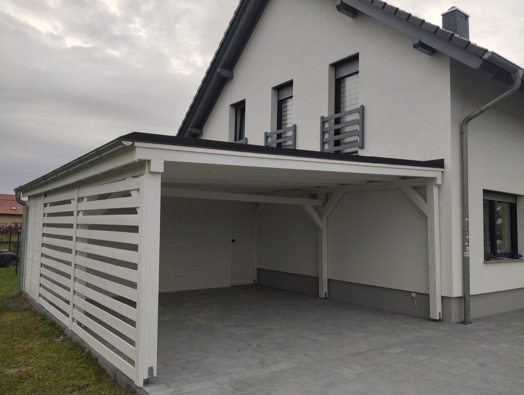 Nowoczesna, biała wiata garażowa z drewnianymi lamelami, przylegająca do budynku z szarymi akcentami. Kostka brukowa przed wiatą. Pochmurne niebo w tle.