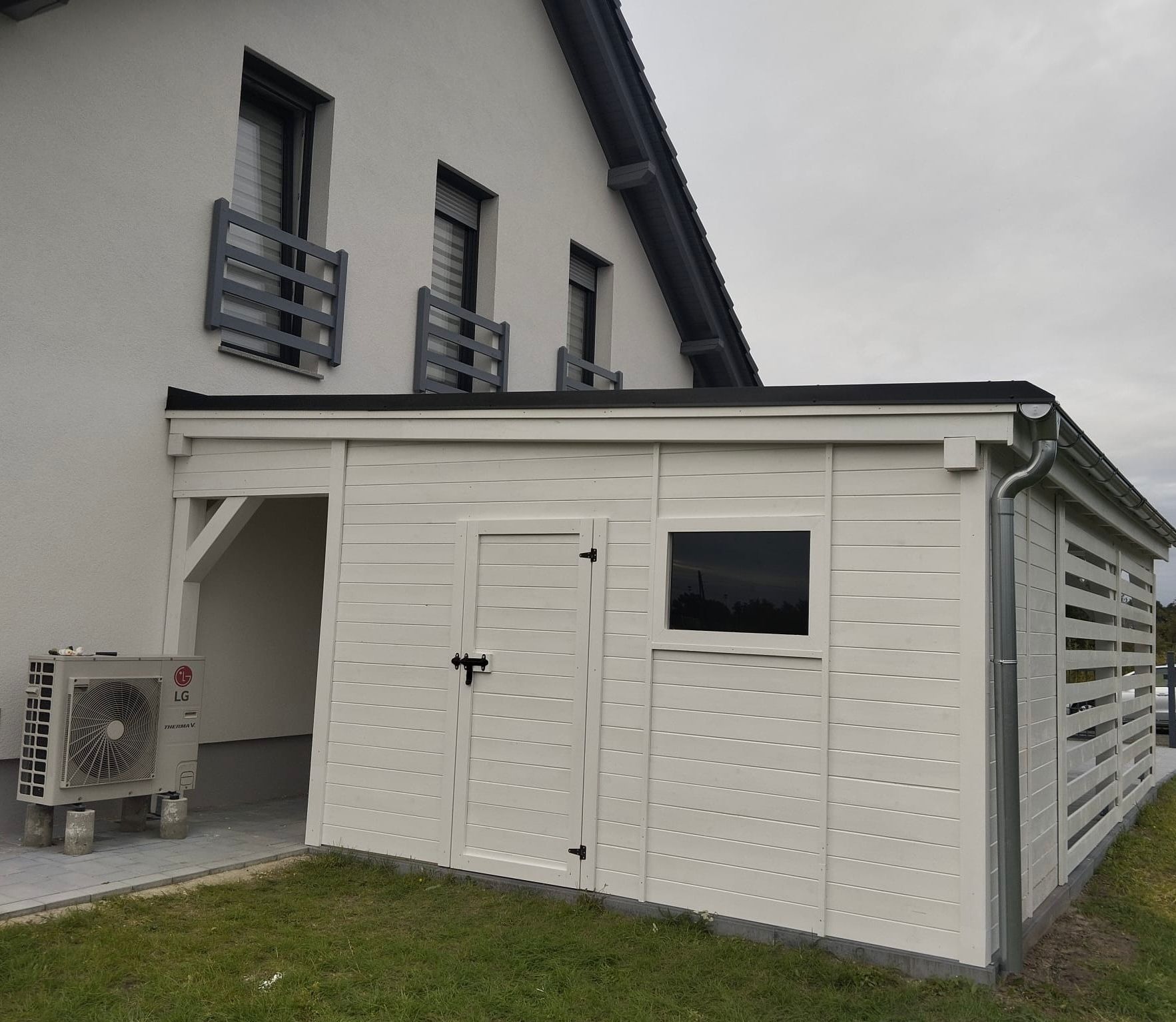 Biała, drewniana konstrukcja przylegająca do domu z klimatyzatorem LG na zewnątrz. Widoczne okno, drzwi z czarnym zamkiem i ażurowa ściana. Dach płaski, szary.